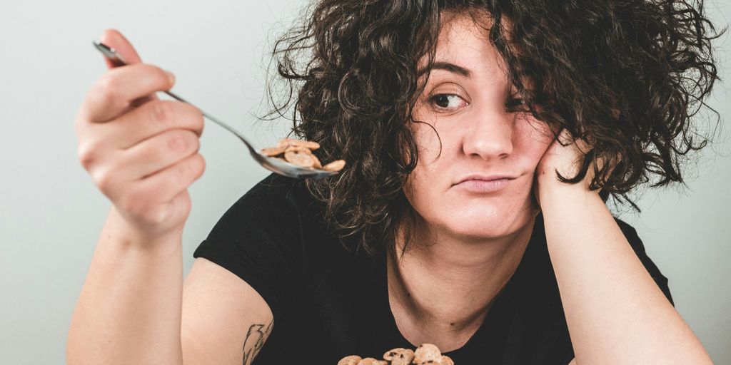 woman with messy hair wearing black crew-neck t-shirt holding spoon with cereals on top