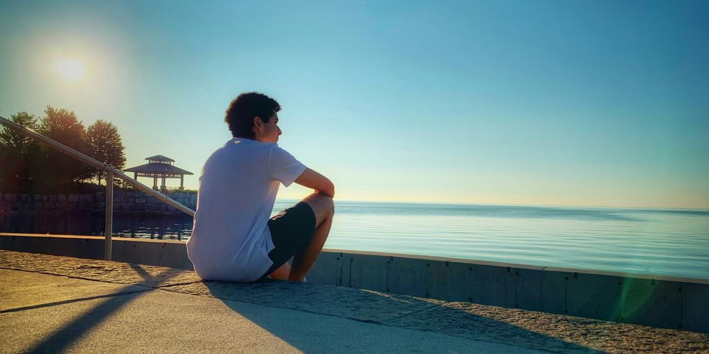 man in white t-shirt sitting on concrete pavement near body of water during daytime