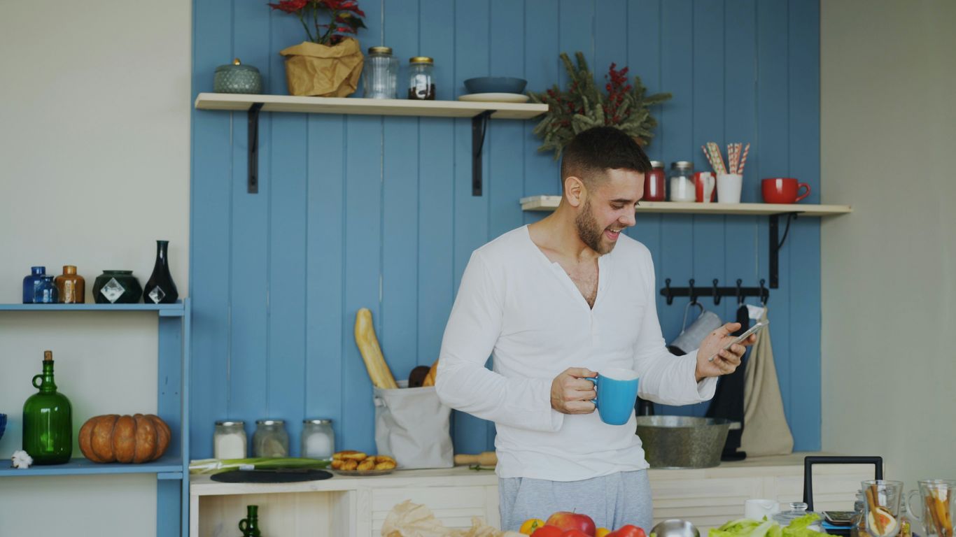 Man holding coffee cup and phone in kitchen.