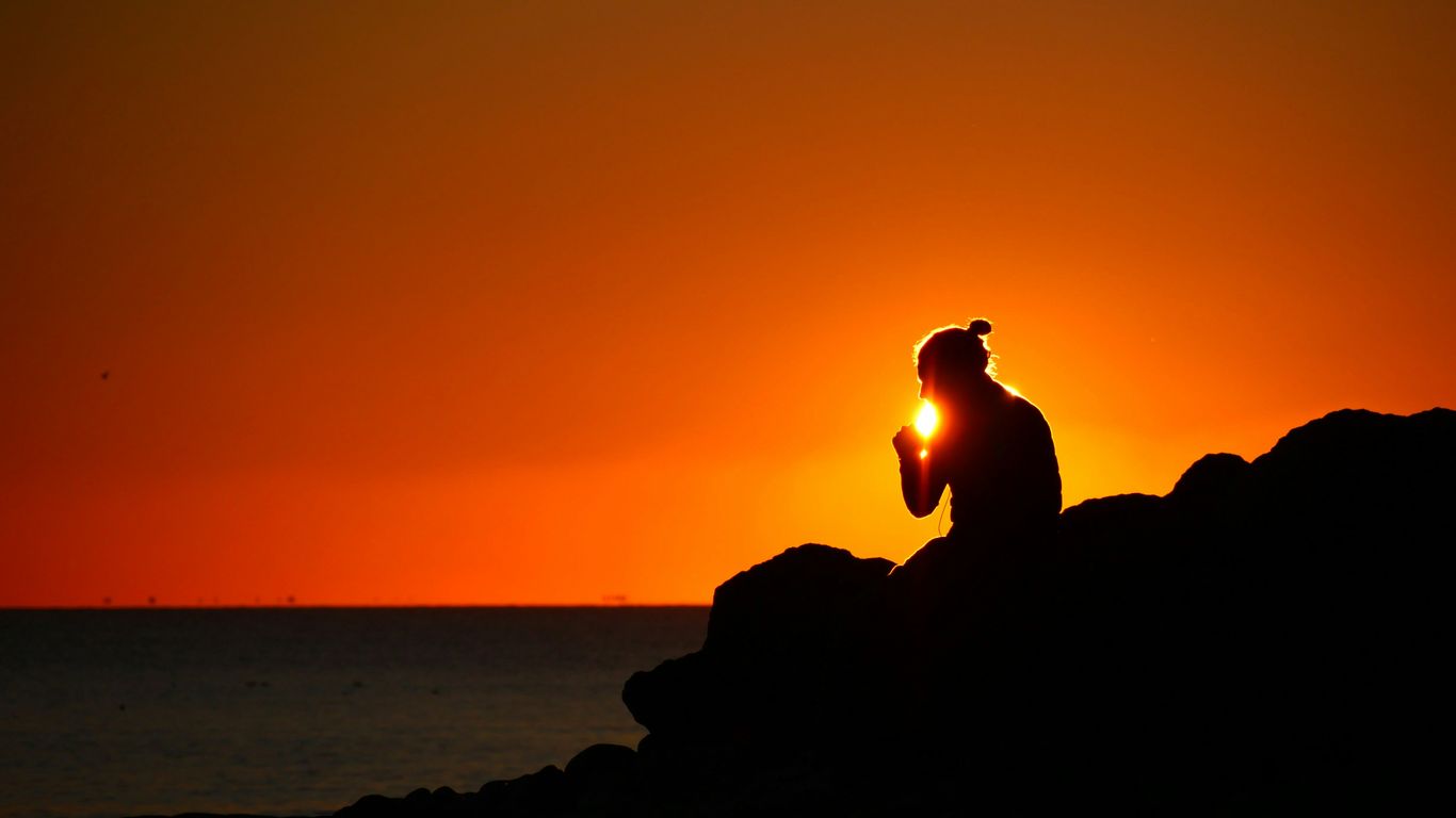 silhouette of person sitting on rock near body of water during sunset